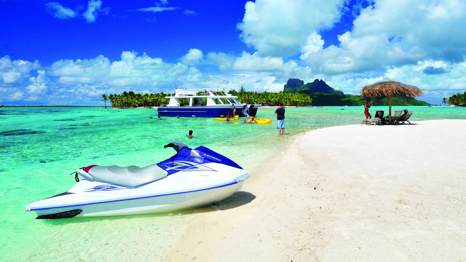 Beached jet ski on the shore, swimmers and paddleboarders by a catamaran in the water. Trees and mountains in the distance.