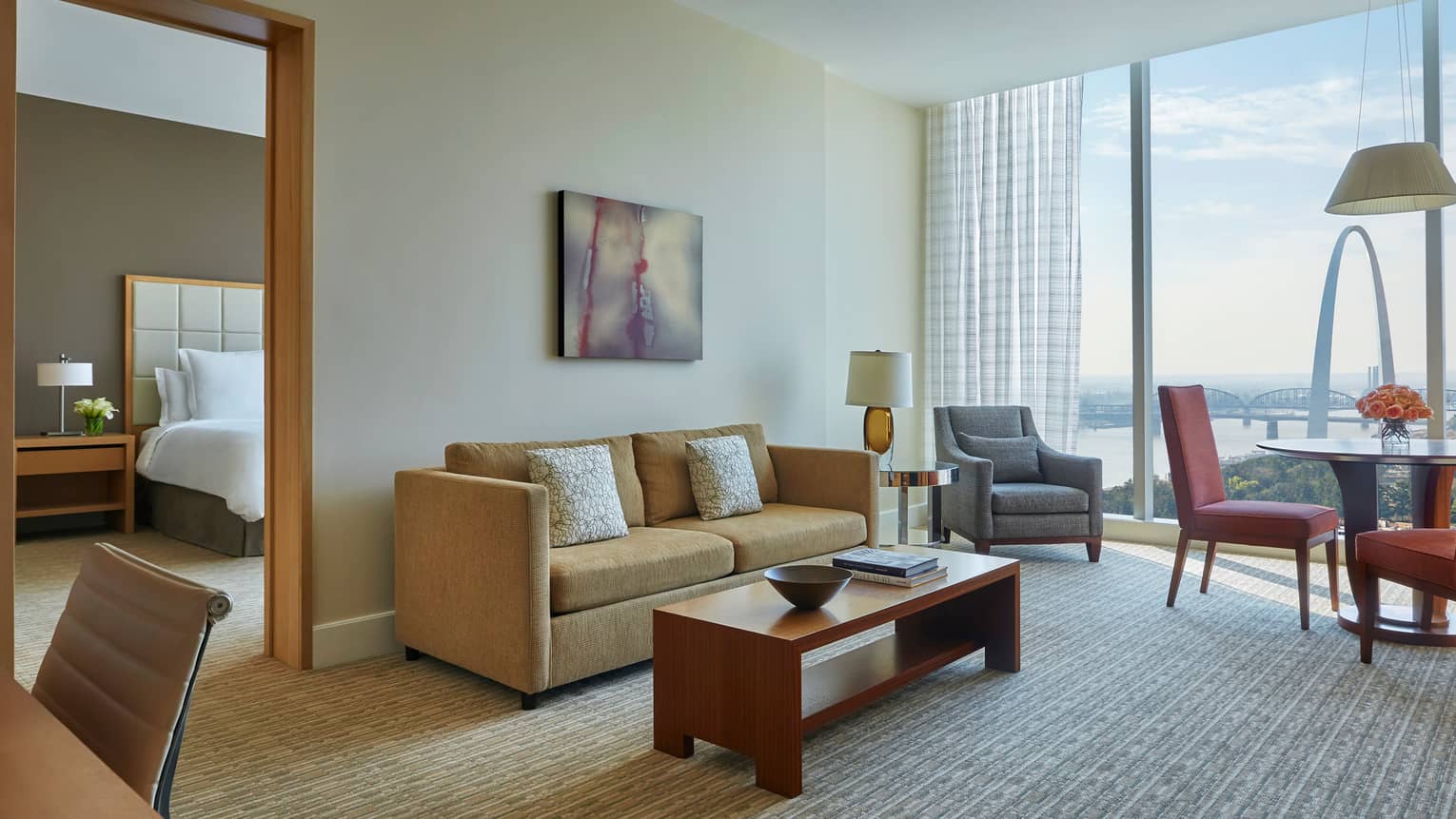 Arch one-bedroom suite living room with gold sofa, grey armchair and dining table, near separate bedroom