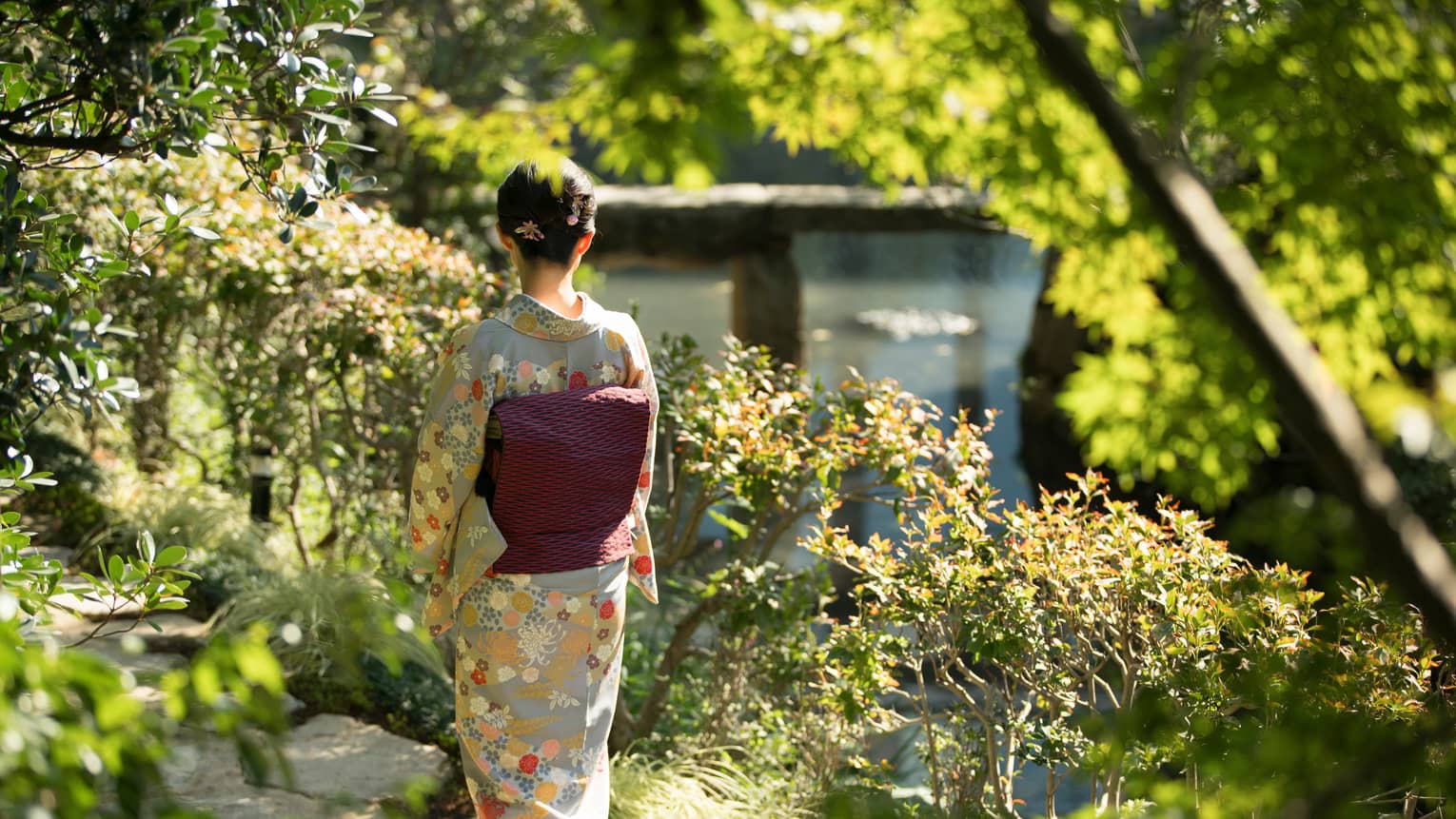 Amid bushes and trees, rear view of a person in a grey kimono with beige and orange flowers and a black and pink obi cushion.