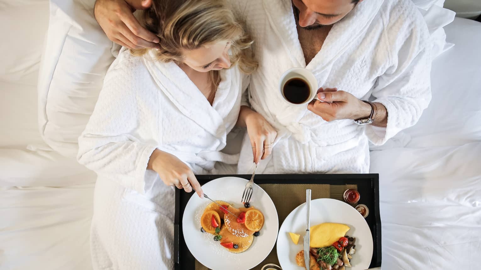Couple wearing white robes cuddle in bed with breakfast tray
