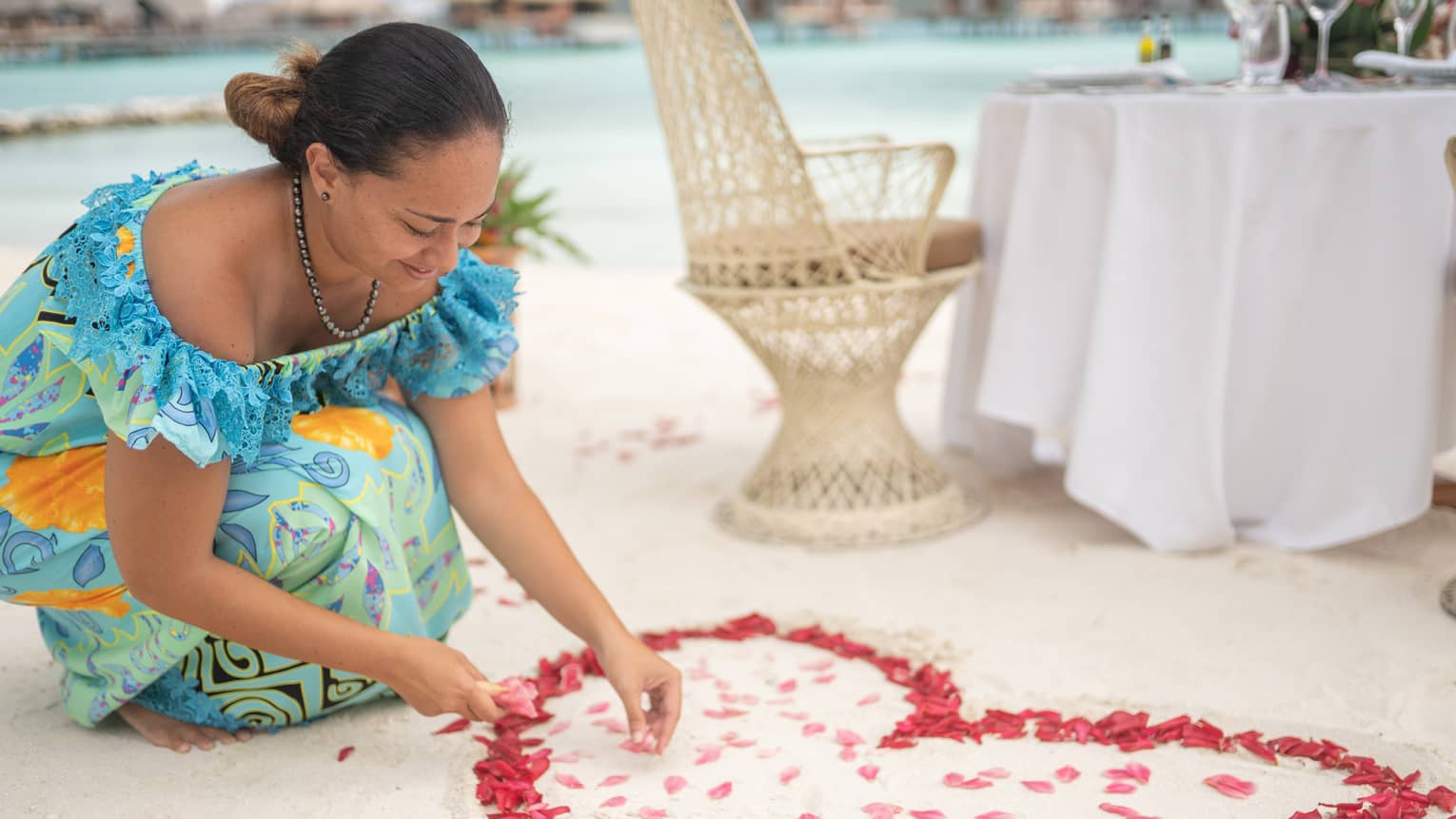 Woman arranges red rose petals into heart shape on sand beach