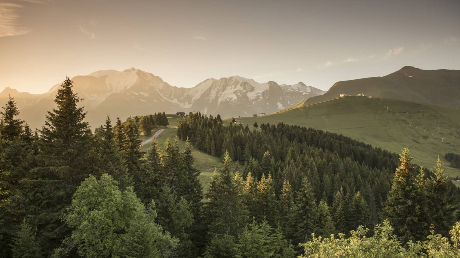 Sun sets over snow-capped mountains, green forests, trails in French Alps
