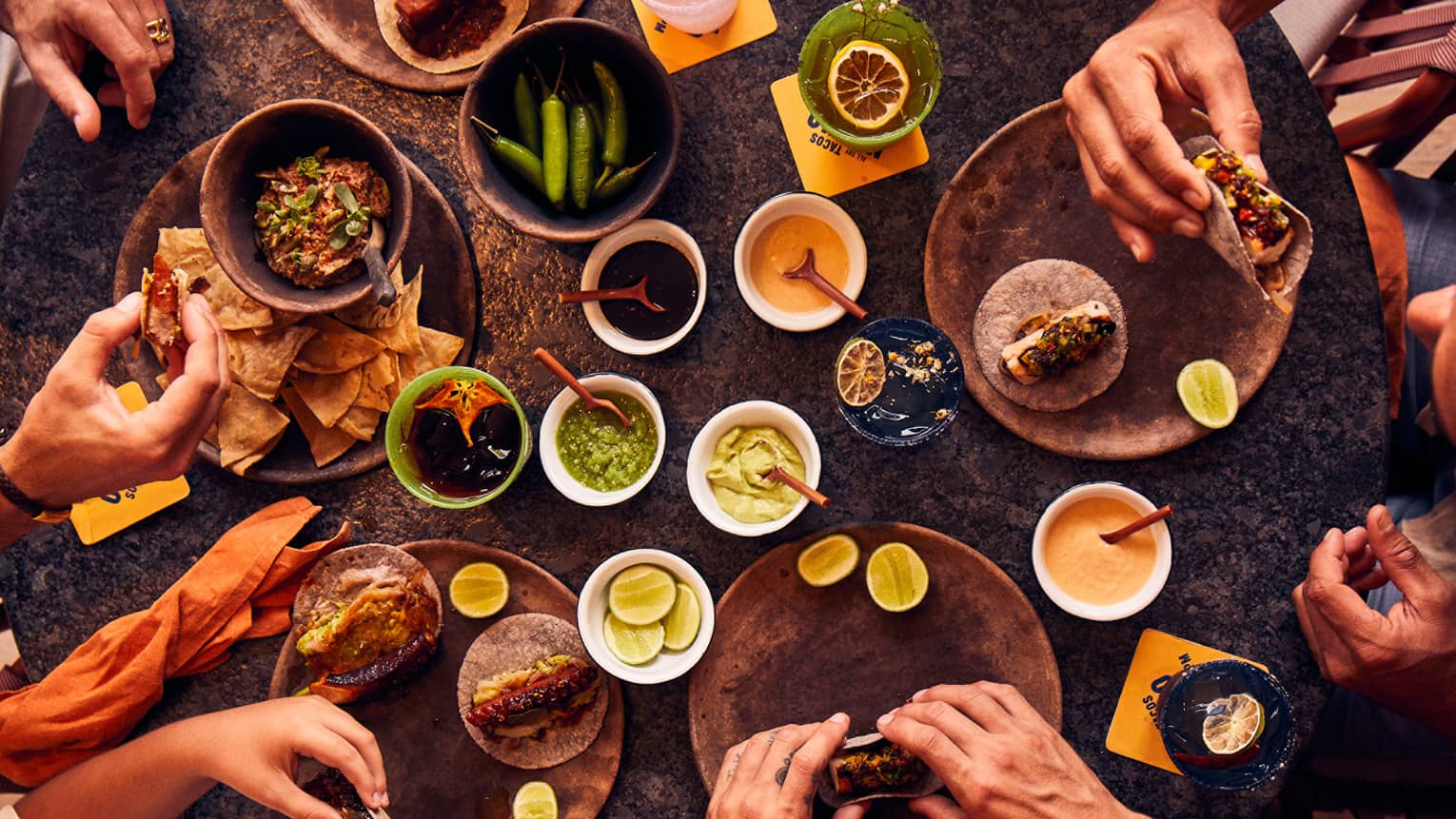 An overhead view of five pairs of hands holding tacos next to bowls of chips, salsa, peppers, limes and sauces.