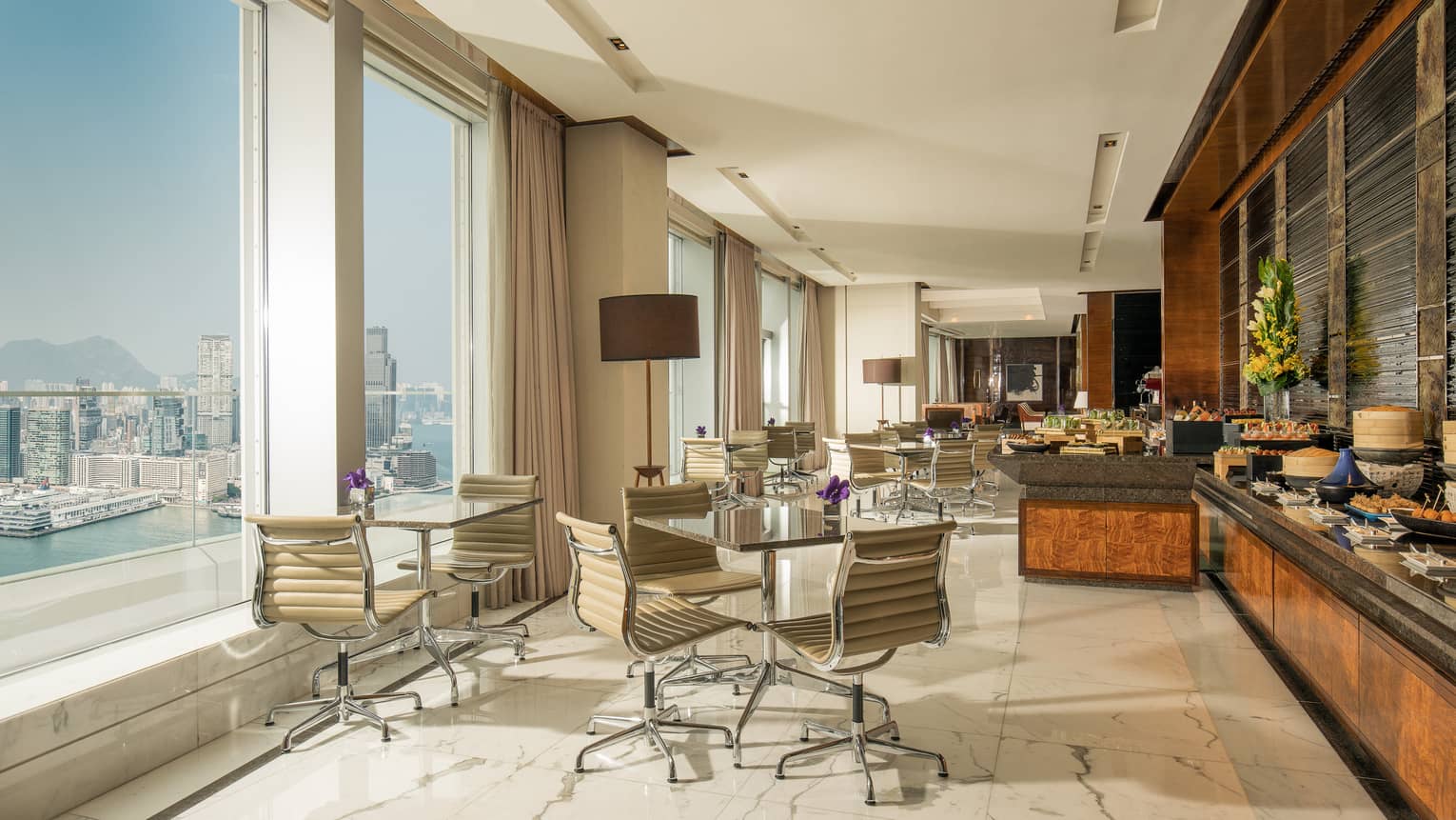 Hotel lounge with wheeled leather desk chairs at round tables across from buffet and beside tall windows with water view