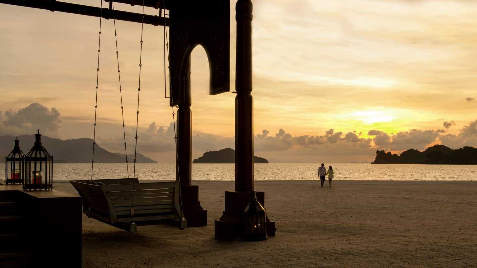 Silhouettes of couple walking along beach past wood swing, pavillion at sunset