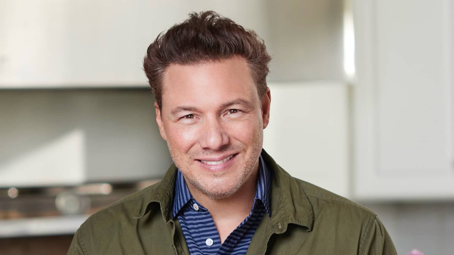 Rocco DiSpirito wearing a blue and white shirt with a green jacket and smiling with their hands clasped in front of them.