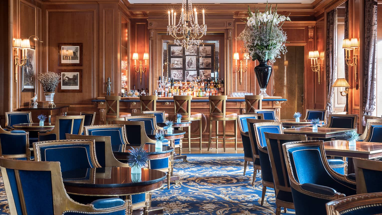 Le Bar des Bergues lounge with elegant blue velvet chairs with wood trims around cocktail tables, crystal chandelier, bar