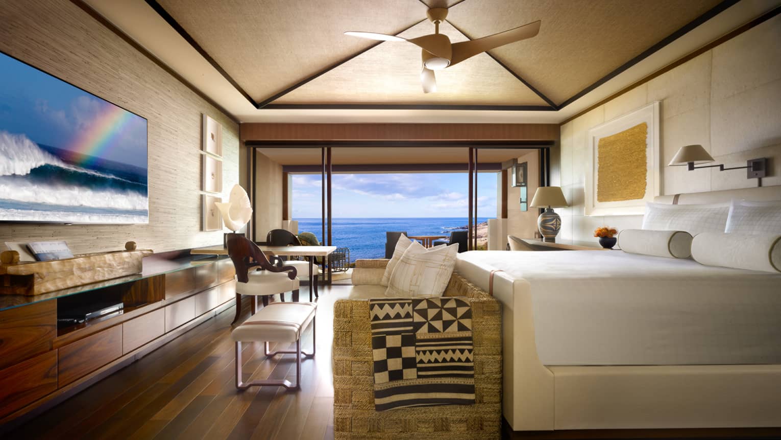 Oceanfront Room bed, small sofa and bench at foot, TV screen displaying ocean waves, rainbow