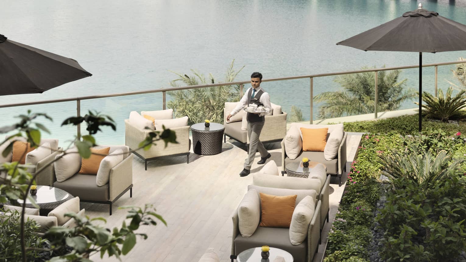 Server in vest and tie carries tray past lounge chairs on sunny rooftop patio with tropical plants 