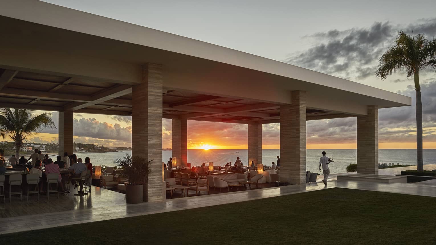 Open-air seaside restaurant with guests dining at sunset, overlooking the ocean.