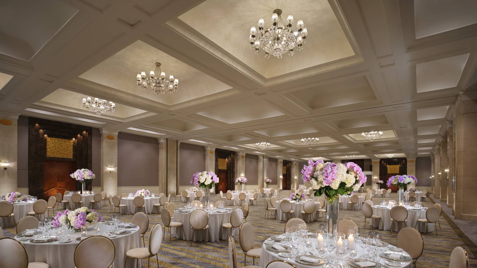 A chandelier-clad ballroom set with several circular tables boasting tall displays of hydrangeas. 