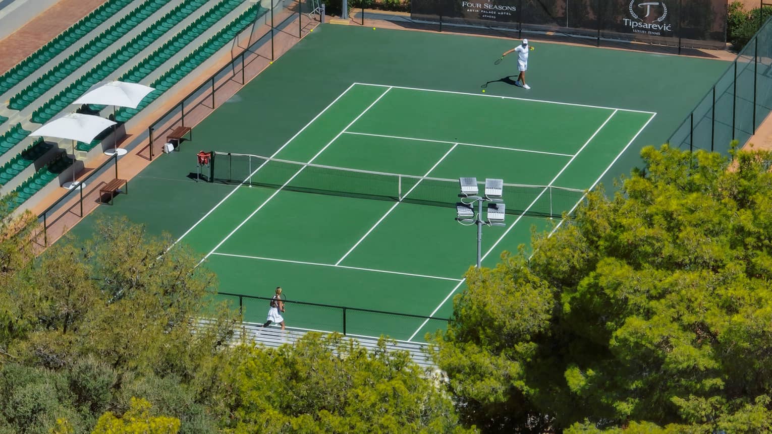 Aerial view of two people playing tennis on court flanked by dense greenery