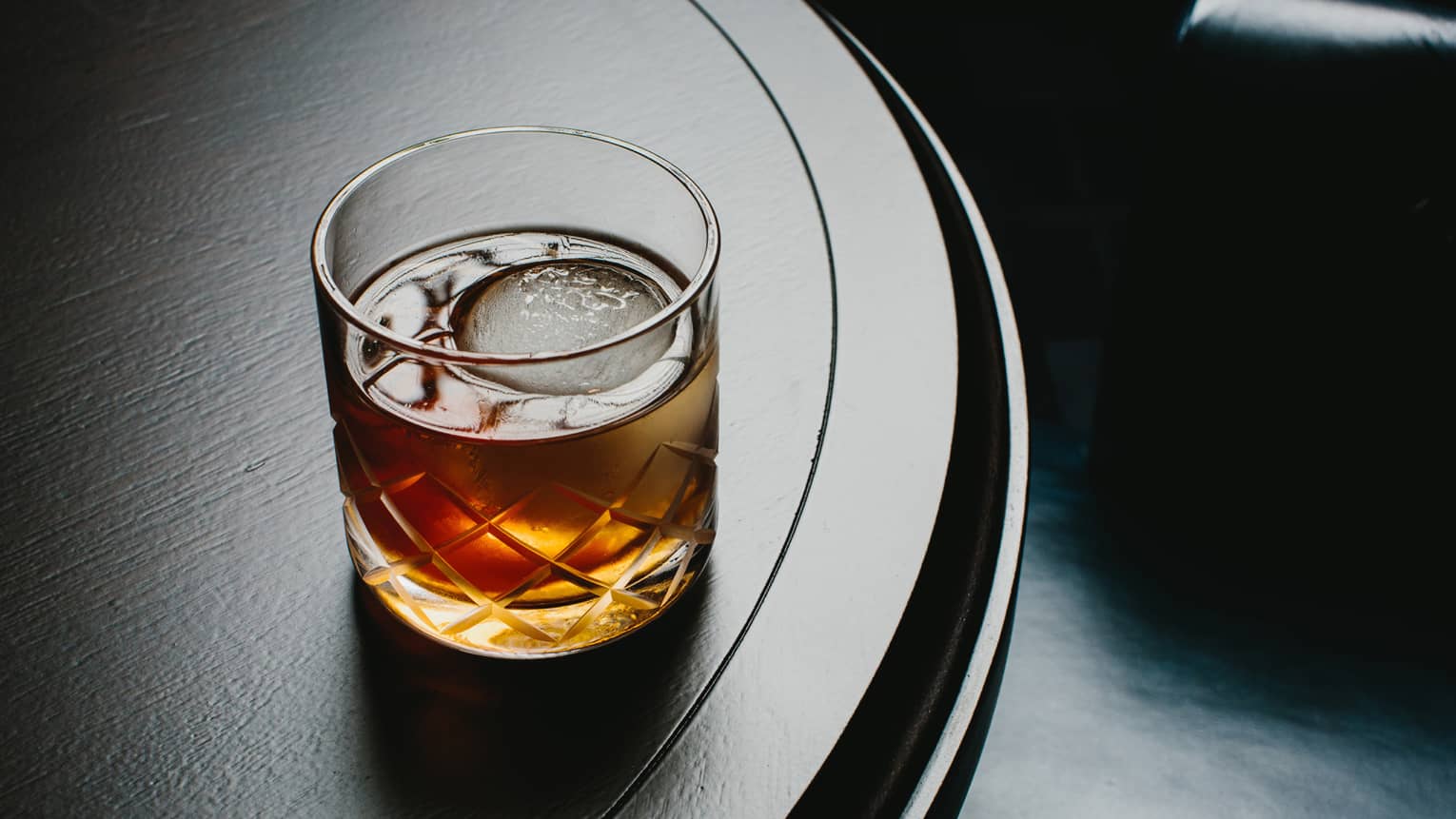 Liquor with large ice cube in rock glass on dark bar