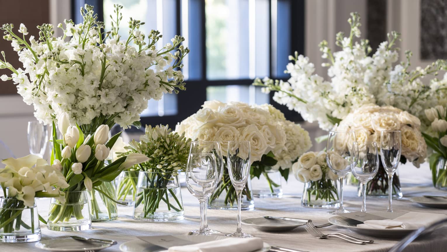 A grey table with an assortment of white flowers in a row, along with white plates and wine glasses.
