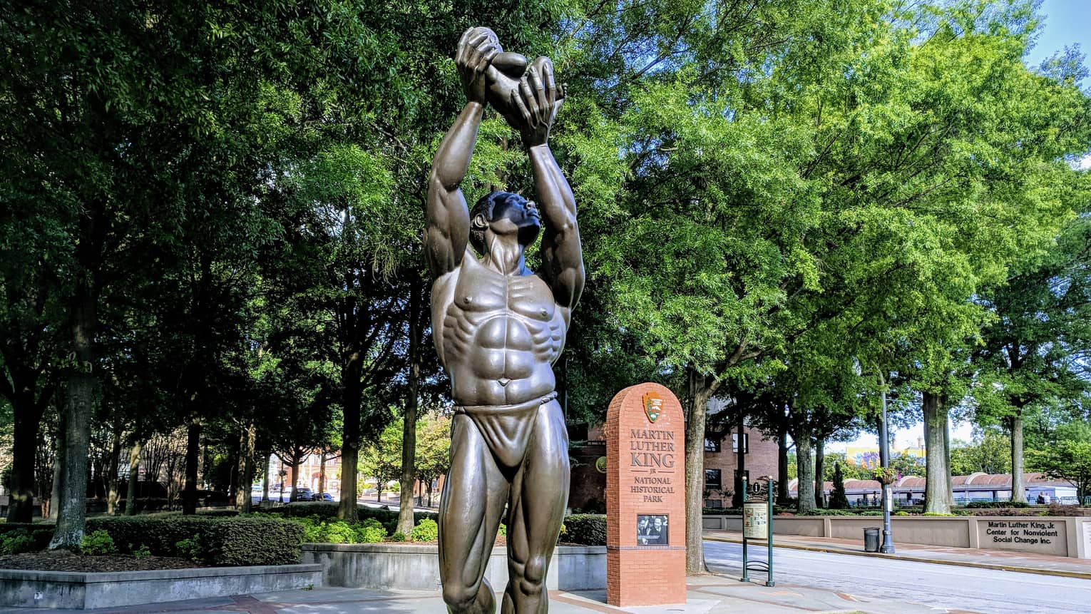 A statue of a man holding something in the air placed in a park with green trees surrounding it.