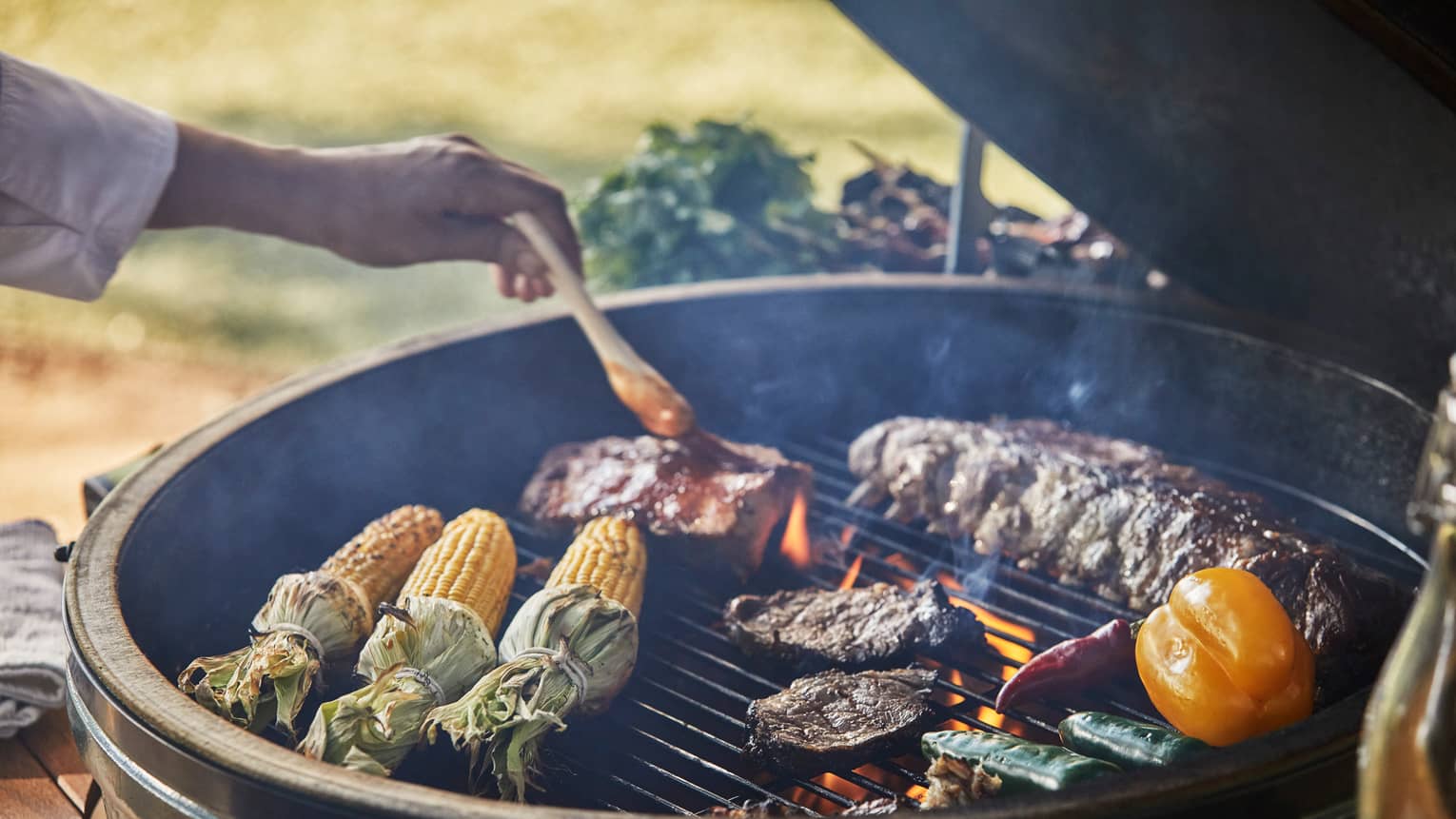 Close-up of chef brushing sauce on ribs on outdoor barbecue with corn, steaks, vegetables