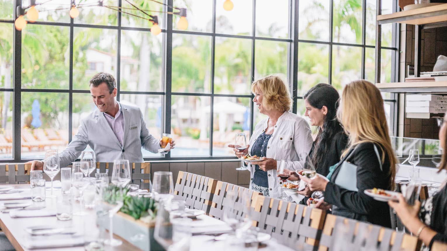 Three women smile, hold wine and appetizers near smiling man with cocktail around long dining table at sunny Seasons Restaurant