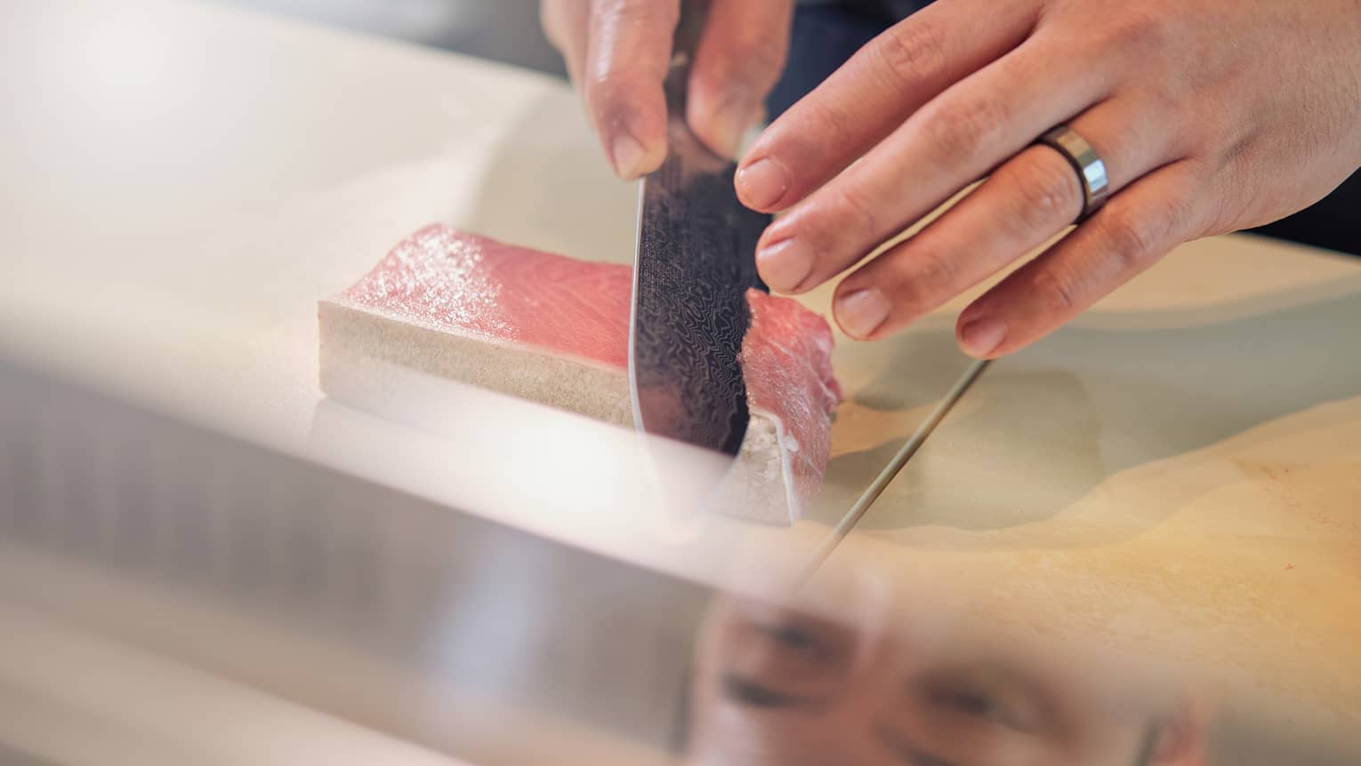 A chef's hands slice tuna on a cutting board behind a glass display which reflects a look of concentration in their eyes.
