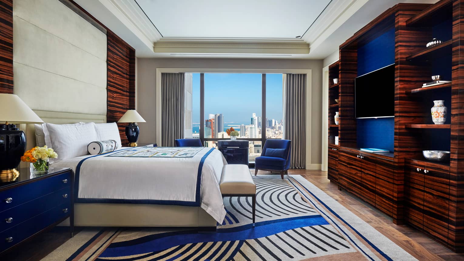 Superior Room side view of bed with decorative blanket and rolled pillow, modern blue carpet, armchair by window