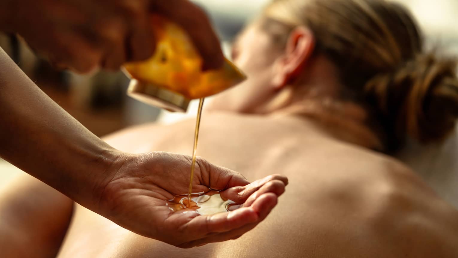 Person pouring oil into their hands while other person lies on spa table.