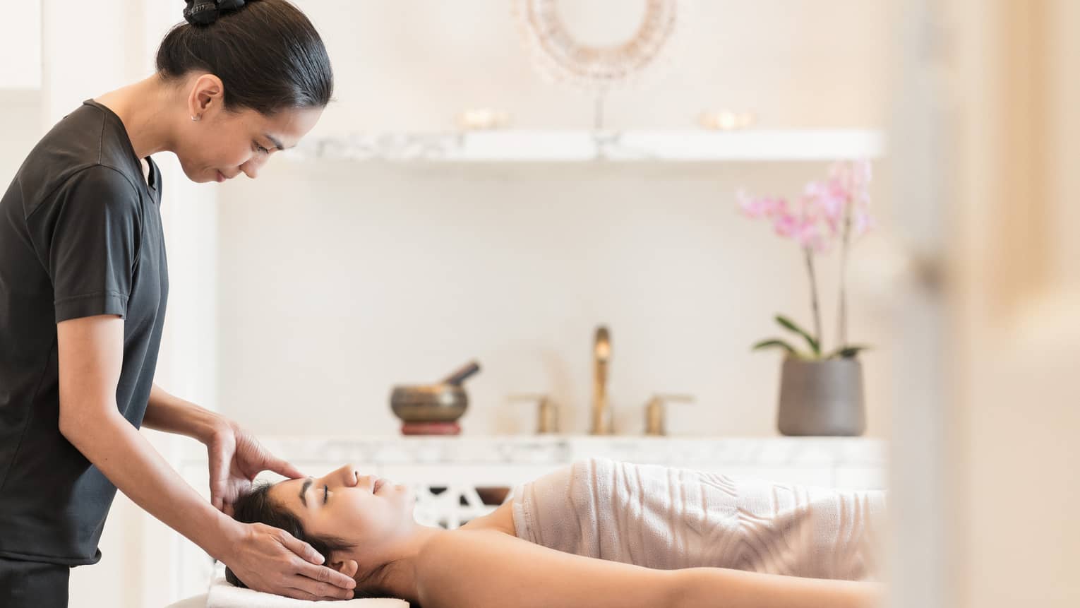 Massage therapist places hands on side of woman's face as she lays under a sheet on spa table