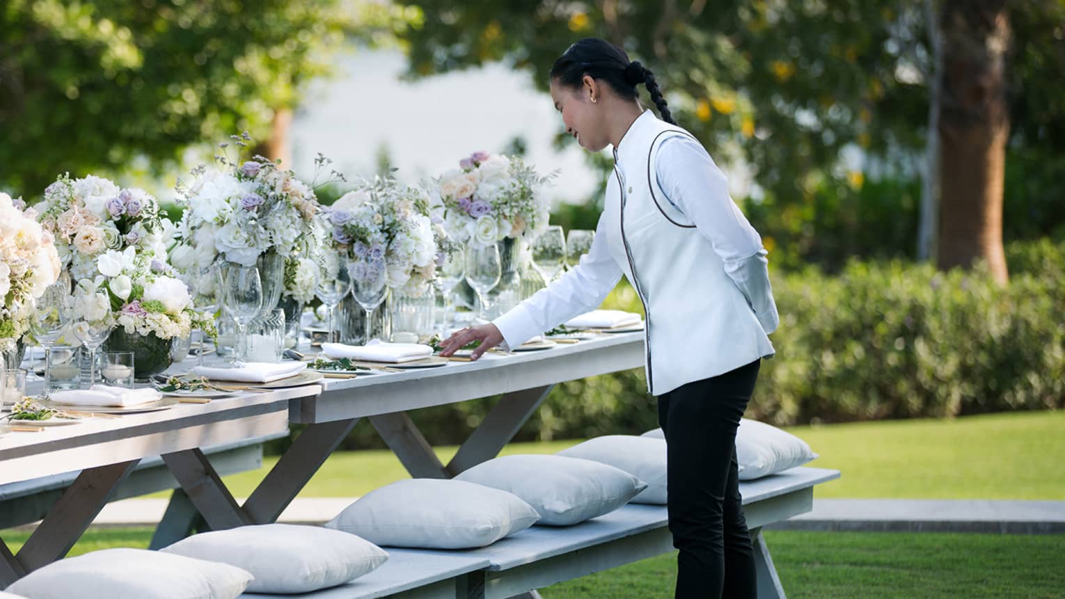 A four seasons staff does the final touches on an outdoor, long banquet table decorated with large white floral arrangements 