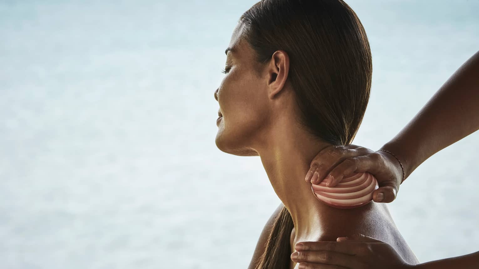 Side close-up of woman from shoulders up with head tilted to side as masseuse rubs seashell down her neck