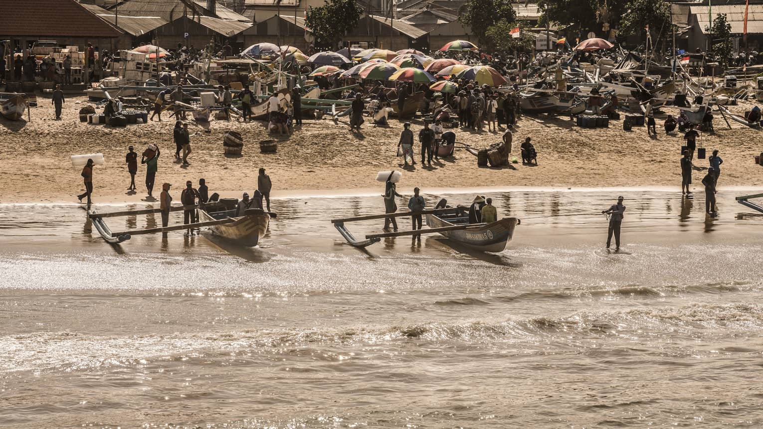 Two outrigger canoes are held upright in shallow water before a beach crowded with people, canoes and colourful umbrellas.
