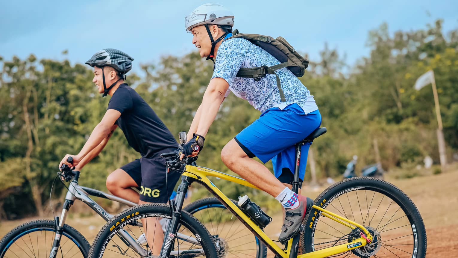 Side view of two smiling mountain bikers in helmets as they coast down a gentle slope on a dirt path alongside trees.
