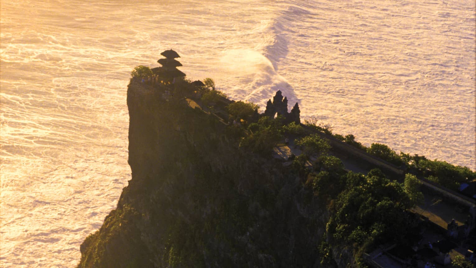 Aerial view of a temple nestled among trees at the edge of a tall, steep cliff; big, white waves crash in the vast sea below.