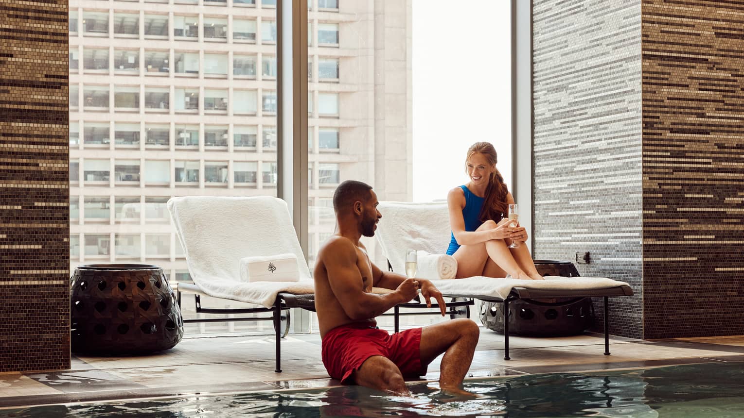 Two guests sitting near the pool with floor-to-ceiling windows behind them.