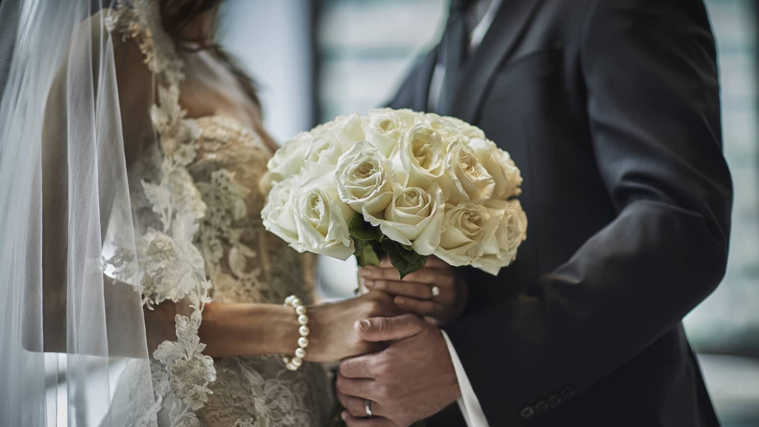 A close up of a bride in a lace gown and a groom in a gray suit hold the bride's bouquet of white roses together.
