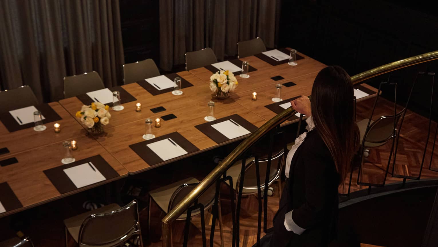 Person wearing a black suit walks down a curved stairwell toward a long rectangular table set for 12