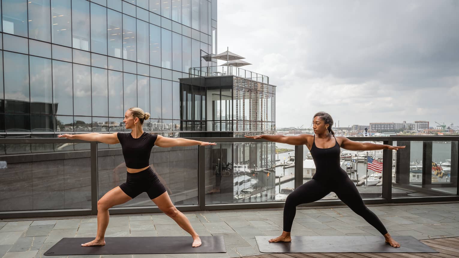 Two people stand in a yoga pose on a rooftop deck, with city views in the background.