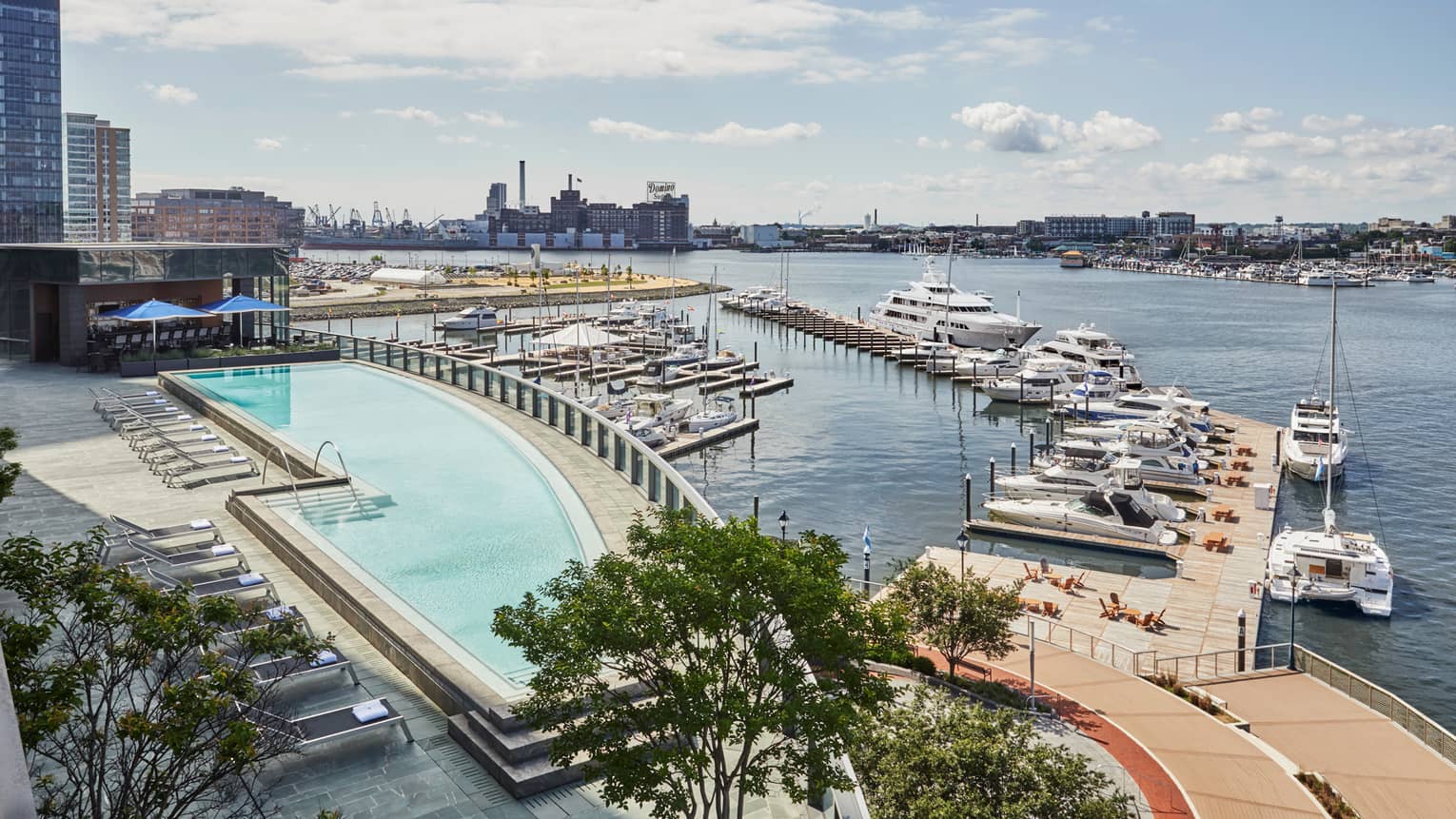 A pool next to a dock with boats, a city can be sen in the distance across the water.