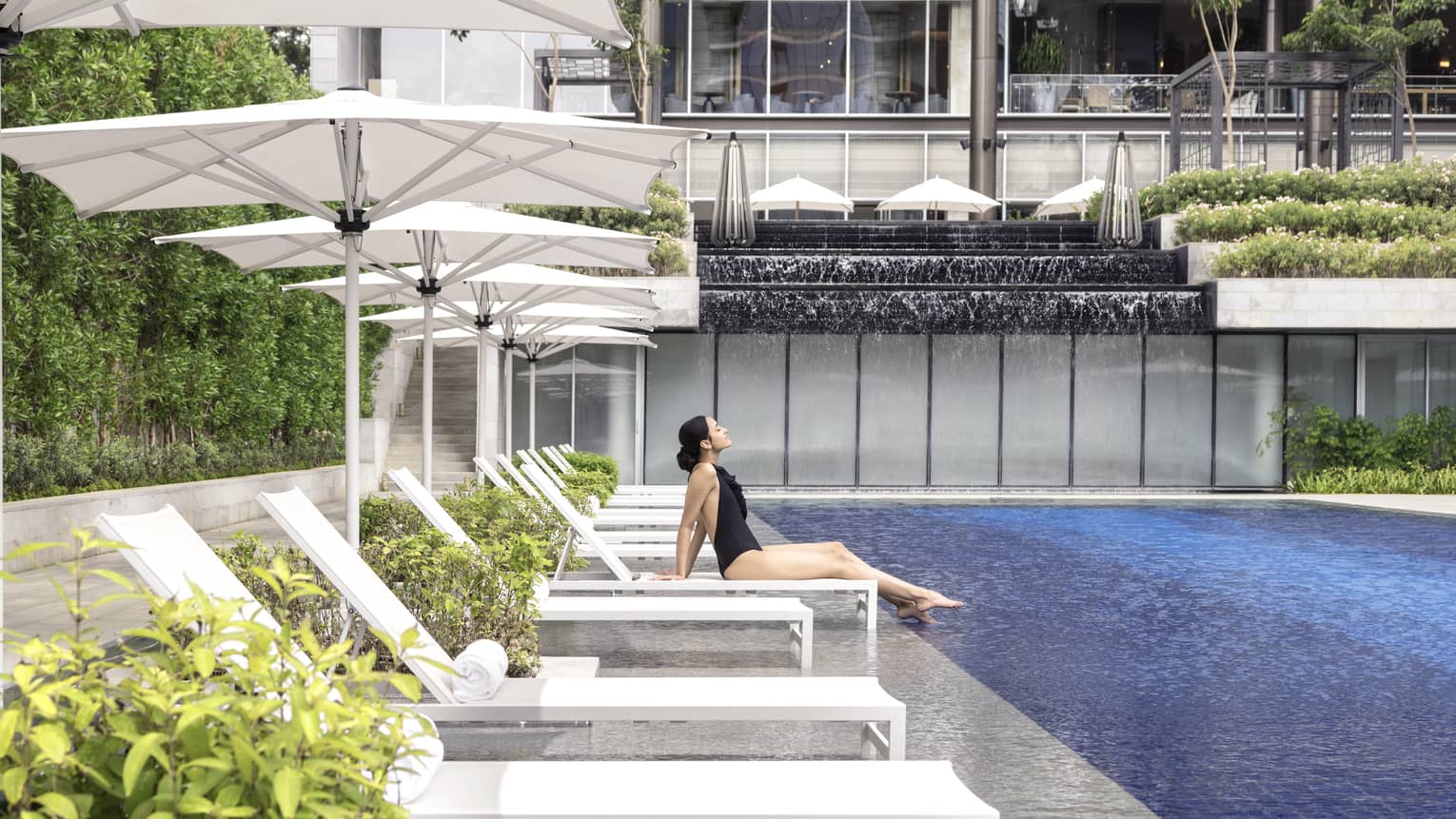 A woman in a black bathing suit relaxes on a chaise by the pool