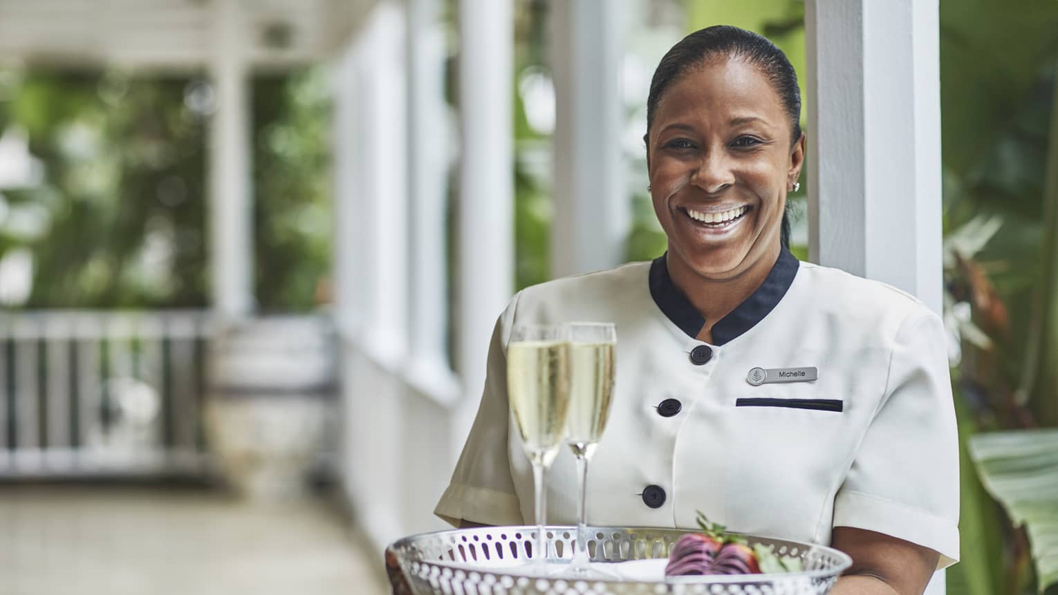Smiling hotel staff holds tray with two glasses of Champagne on veranda