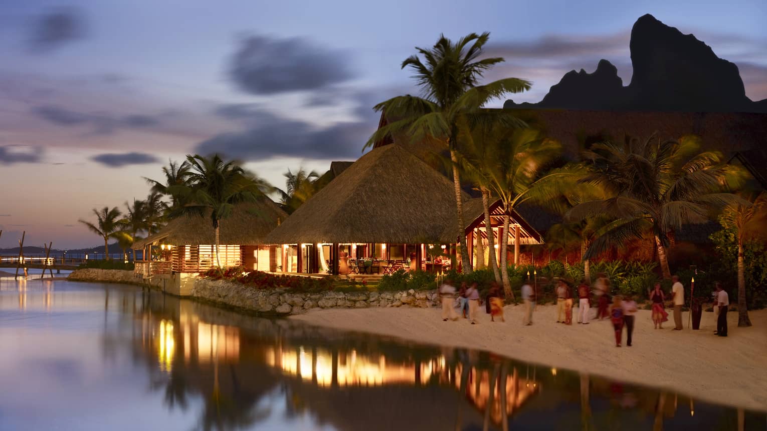 Guests on sand beach by pavilions with lights at night