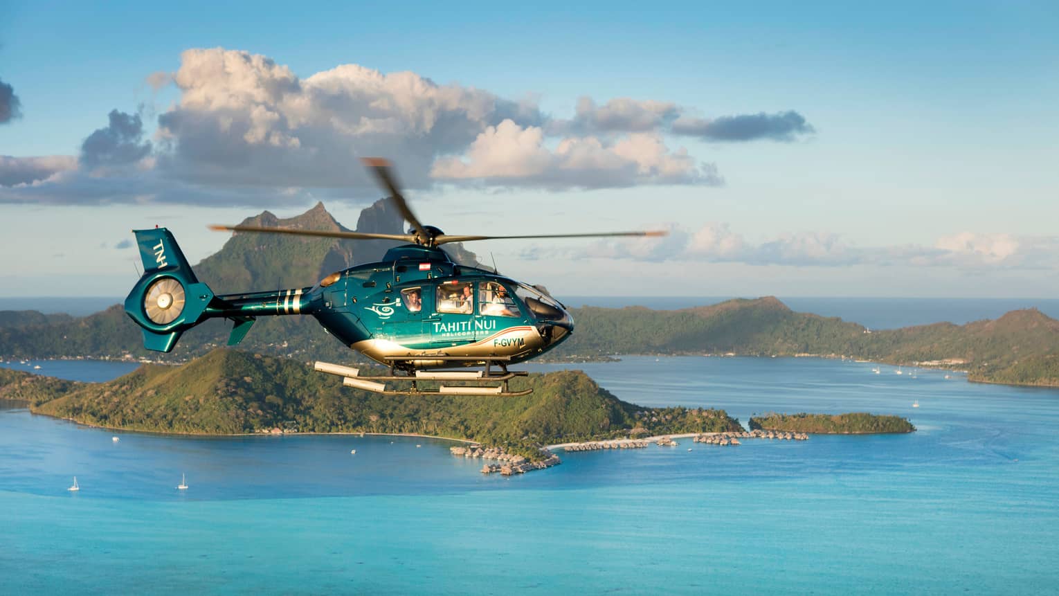 Guests peer out of a helicopter as it flies over turquoise ocean and past forested islands lined with rows of bungalows.