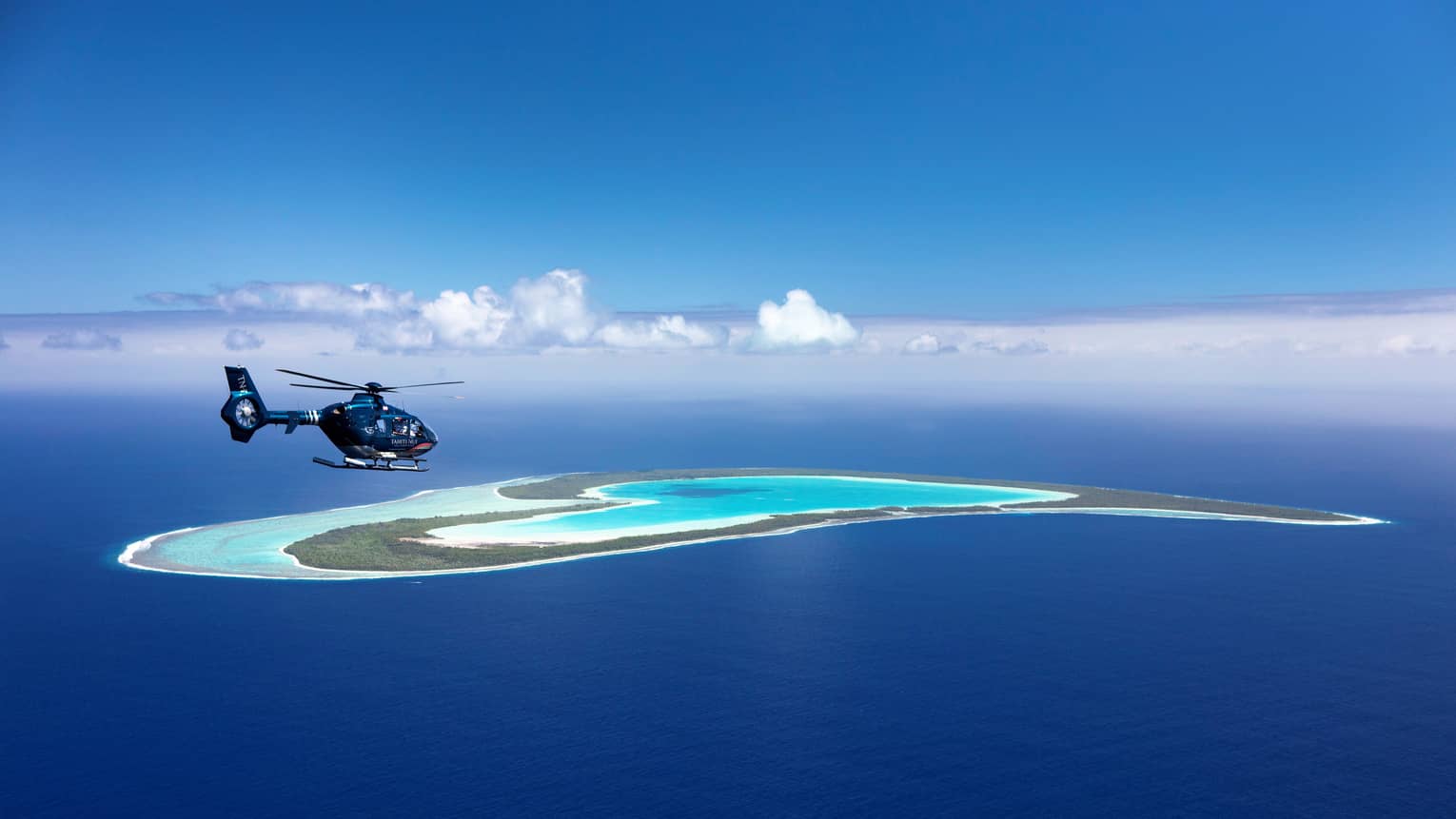 A helicopter flies over a teal lagoon, a heart-shaped island in the distance surrounded by endless ocean and sky.