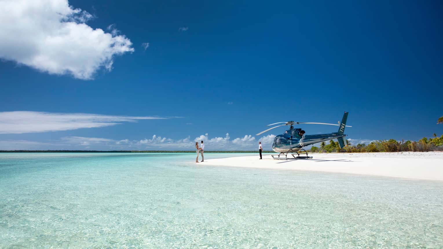 A couple holds hands on a white sand beach beside the pastel blue ocean; a parked helicopter and its pilot wait nearby.