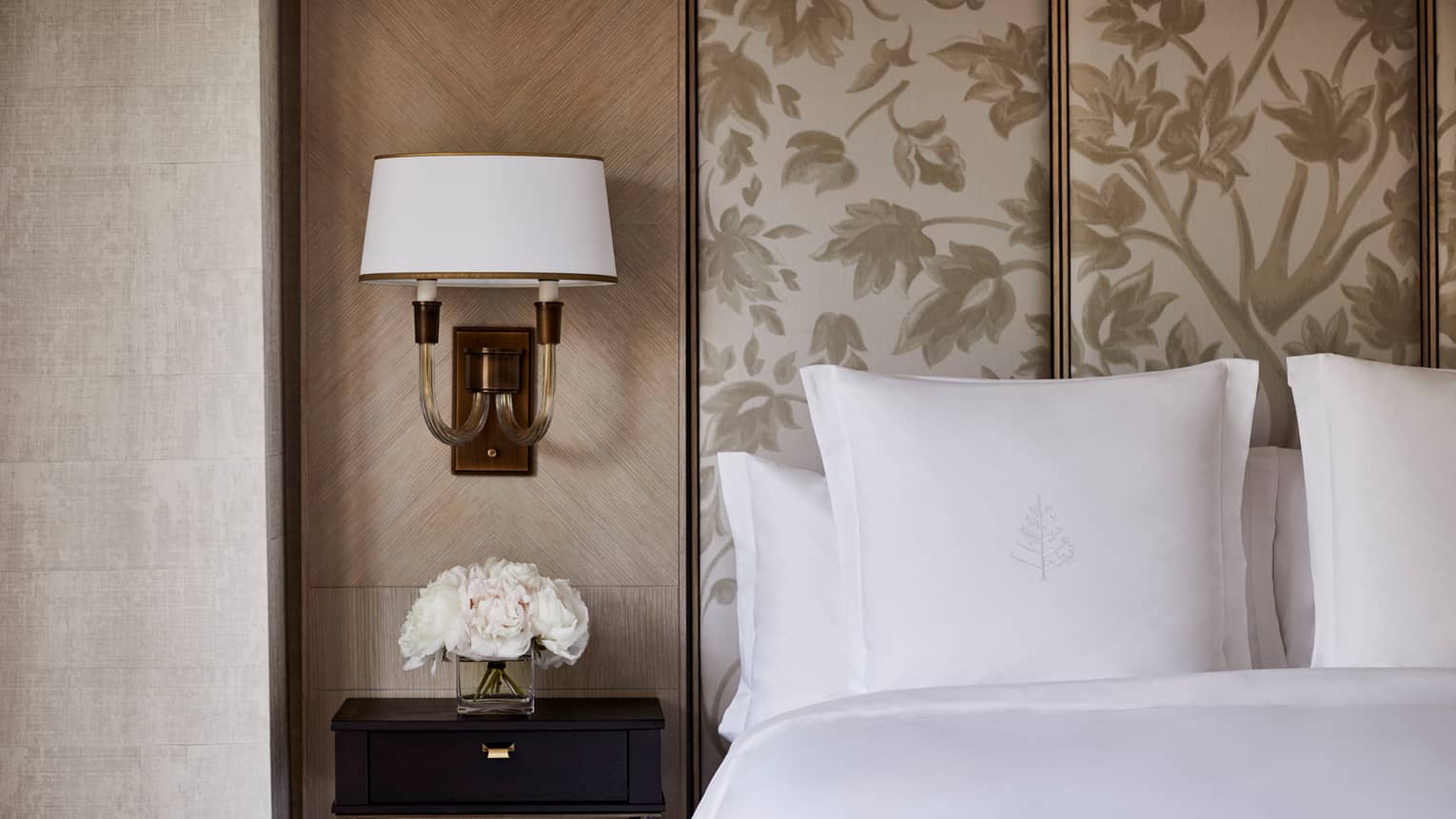Deluxe Accessible Room close-up of bed, white pillows, beige headboard, small reading lamp on wall, white flowers on nightstand