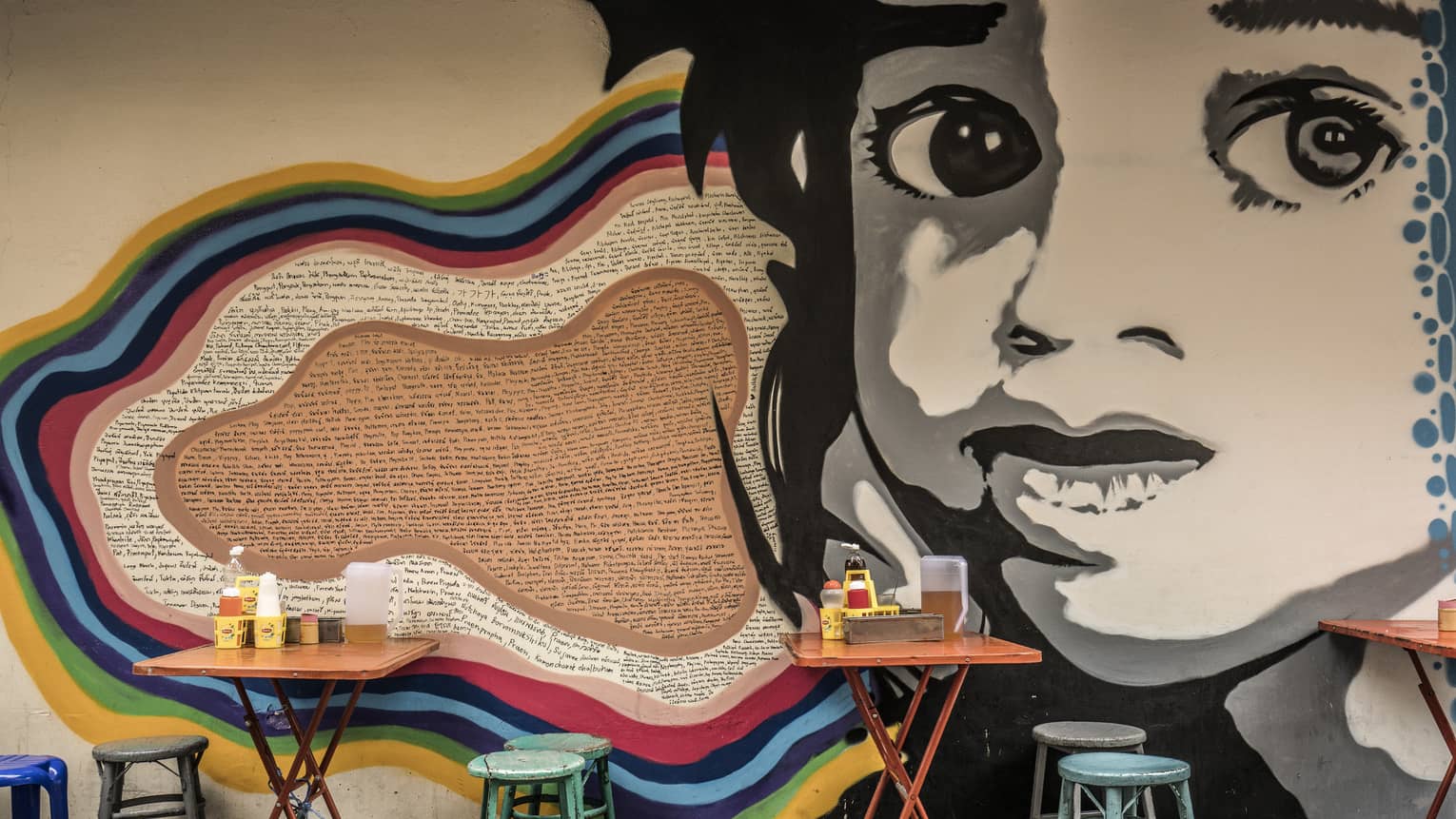 Street art mural featuring a large black-and-white face on a wall, with small, colourful dining tables and stools in front it