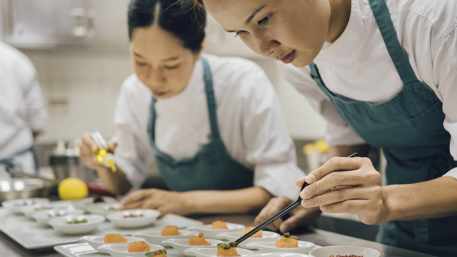 Two chefs in green aprons use kitchen tweezers to place final touches on one-bite dishes in white bowls