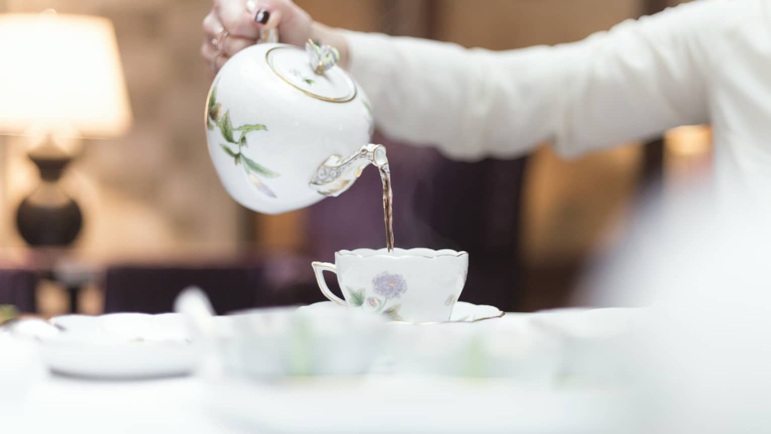 Person holds floral tea pot and pours tea into cup and saucer 