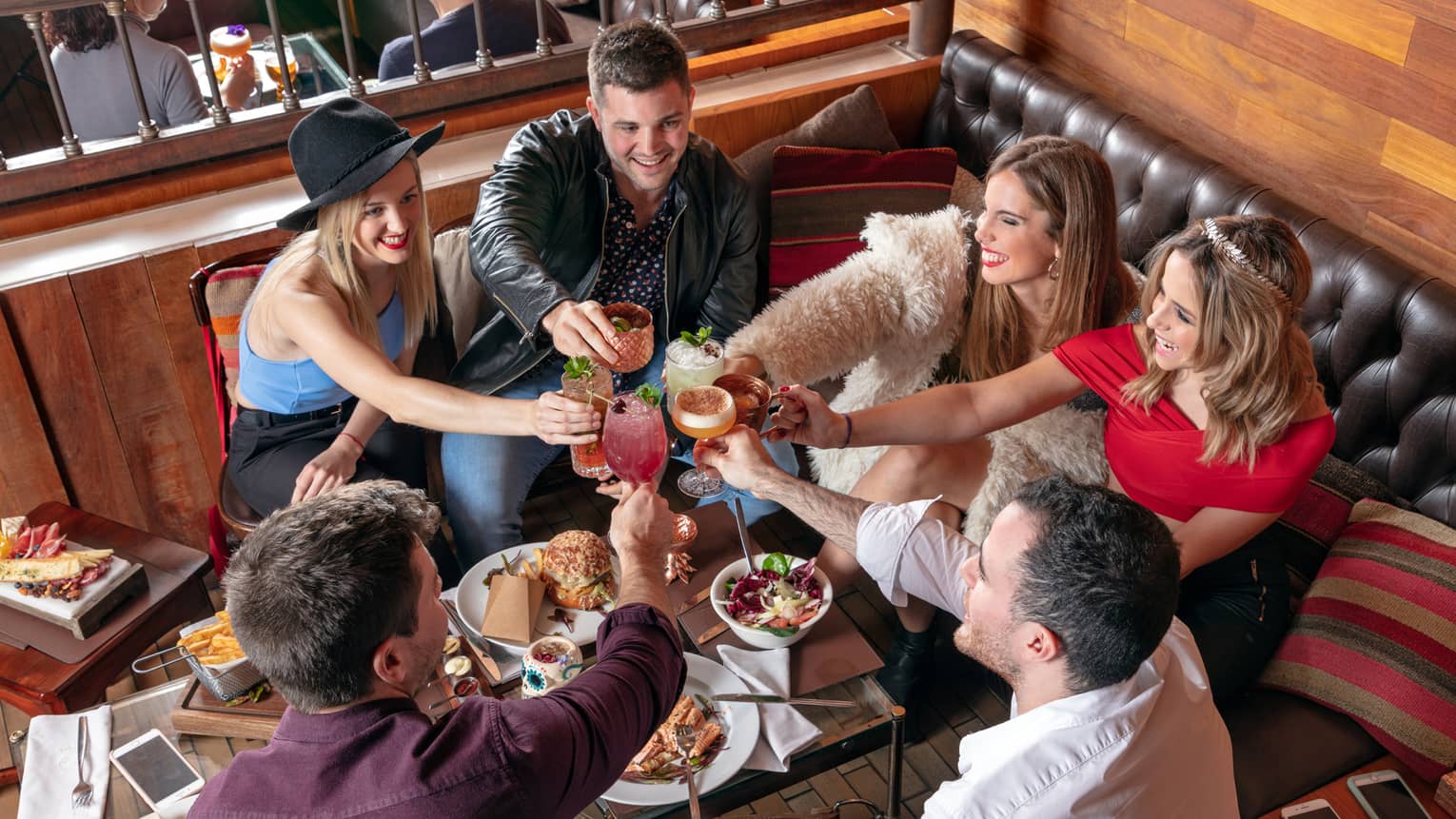 Group of friends sitting in restaurant lounge booth at table with appetizers, toast with cocktails