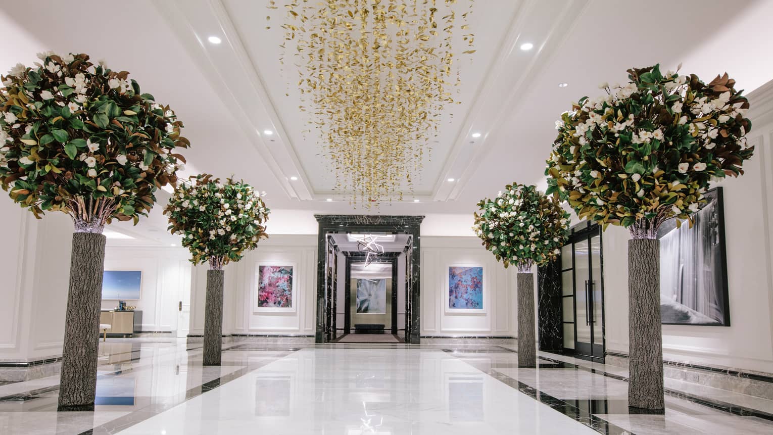 Small trees lining a lobby area with a gold chandelier hanging from the ceiling.
