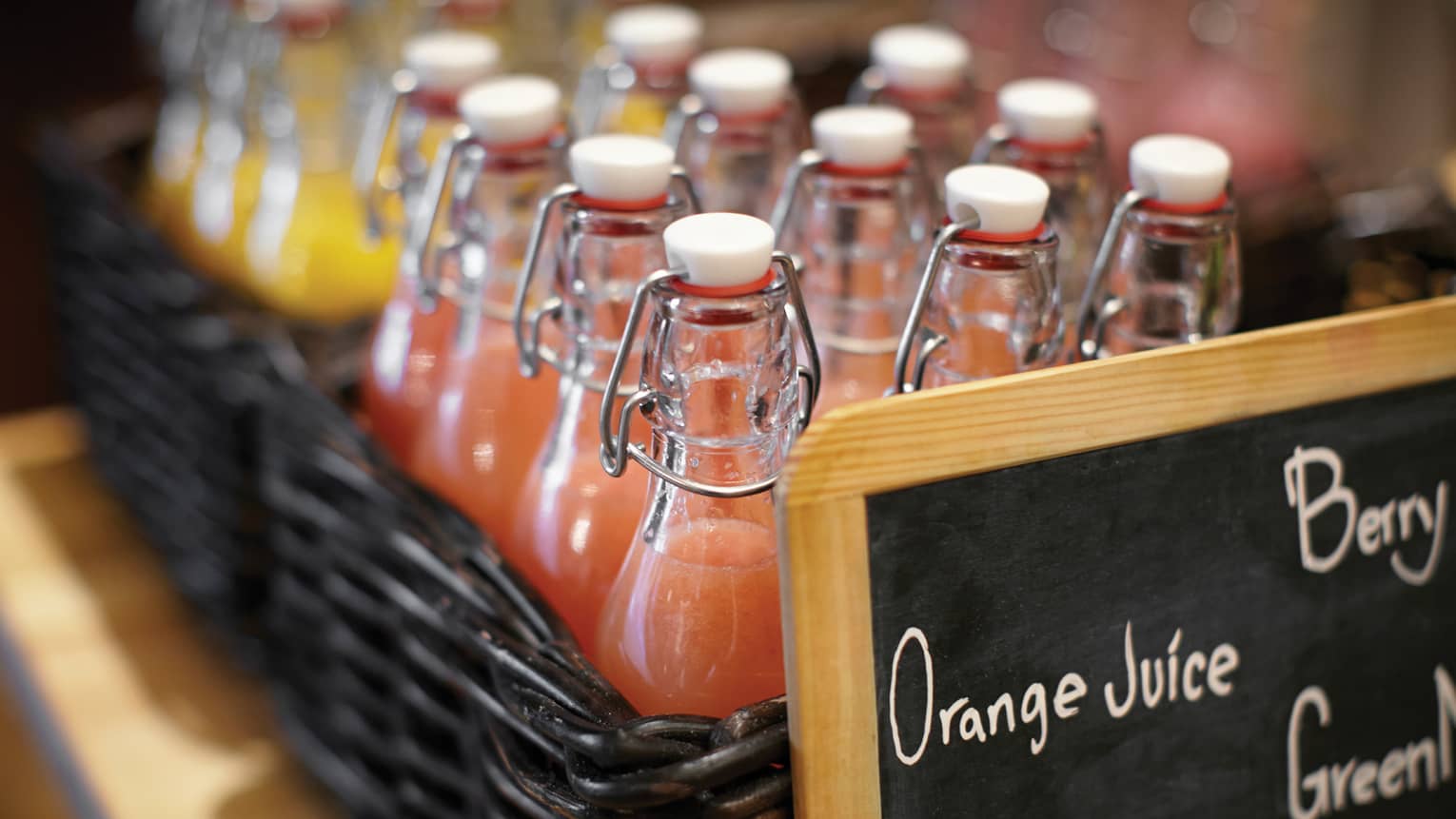 Glass bottles of orange and grapefruit juices in a dark brown wicker basket with a chalkboard sign in front