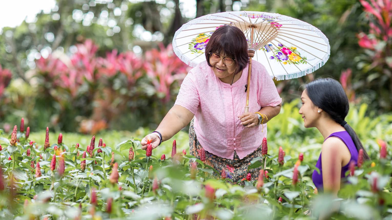 Two women chat in a bed of tropical plants, The woman on the left is holding a floral parasol.