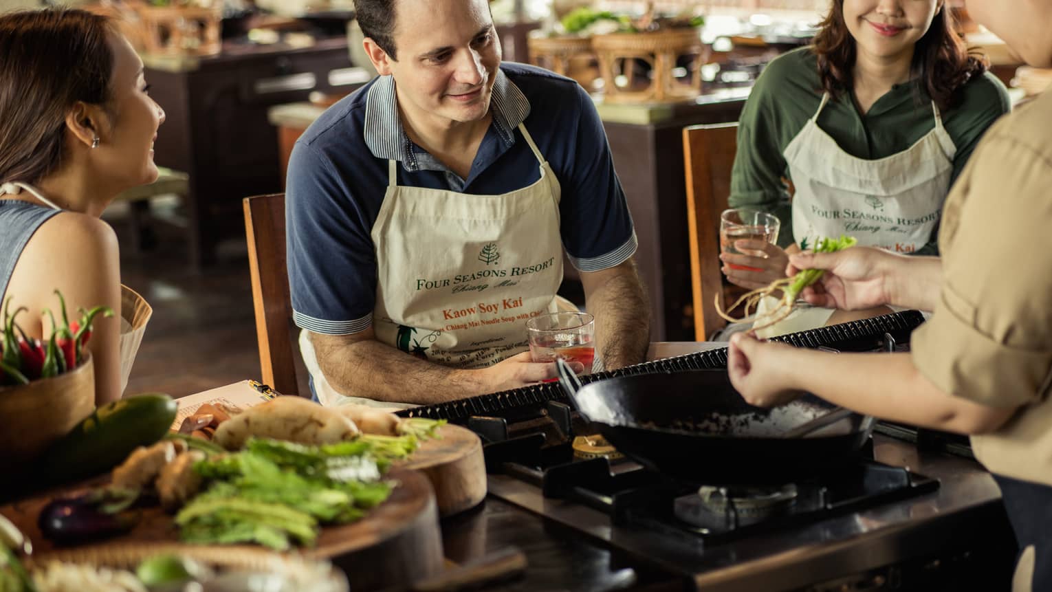 Three guests in aprons sit on stool in front of large wok, chef during Thai cooking class